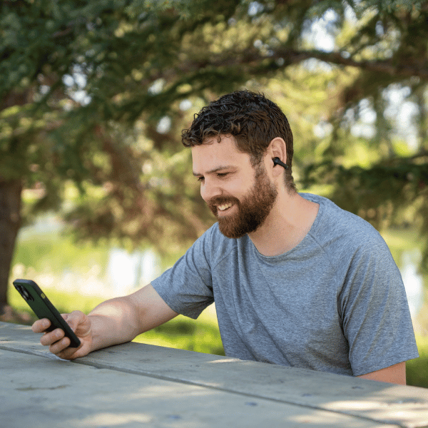Smiling man at an outdoor table using his phone while wearing open-ear earbuds