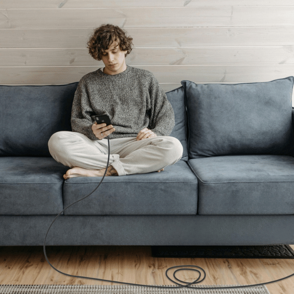 Man sitting cross-legged on a sofa using his phone while it charges on a long cable